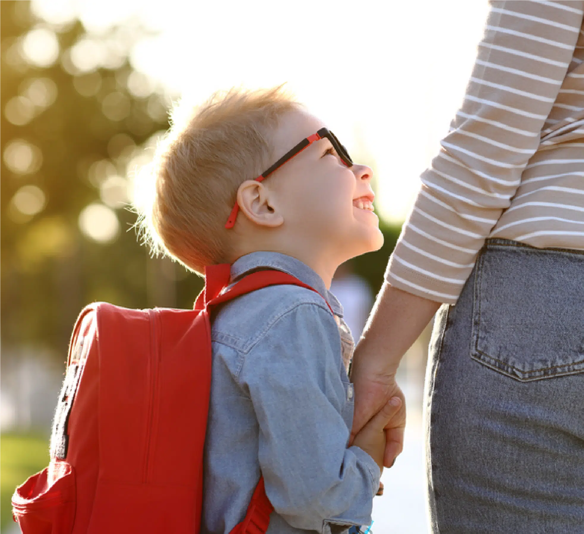 A happy, little boy with glasses and a red backpack looking up at the person holding his hand.