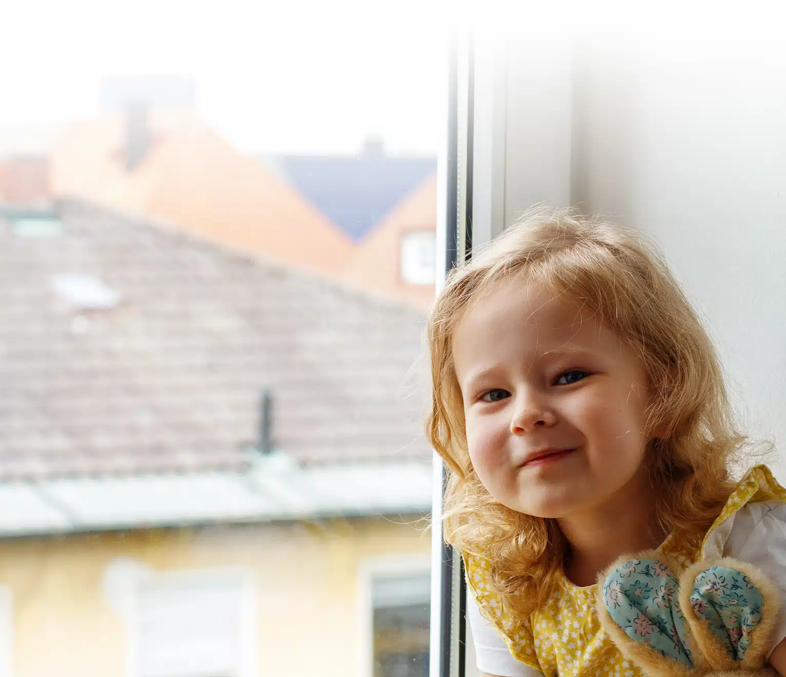 Image of a cute little girl smiling at the camera, sitting by a window, holding a little stuffed bunny.