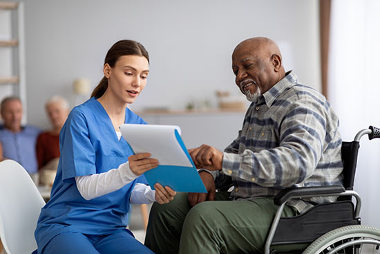 Young woman in blue workwear nurse helping senior black man in wheelchair with questionnaire, african american pensioneer filling papers at nursing home, having assistance
